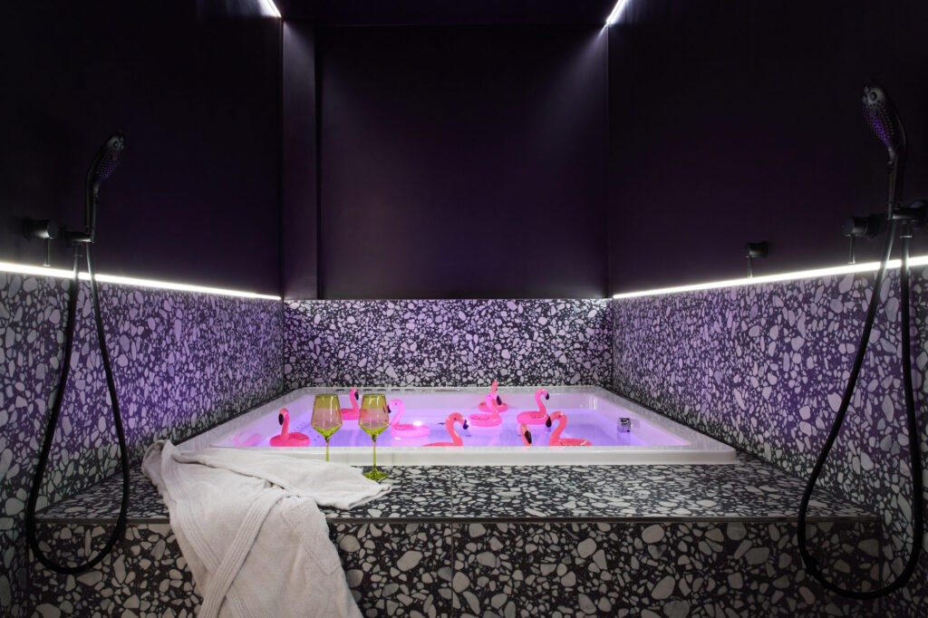 A playful view of the premium jacuzzi at The Circle Berlin private spa, decorated with 10 pink plastic flamingos, a luxury bathrobe, and elegant wine glasses.