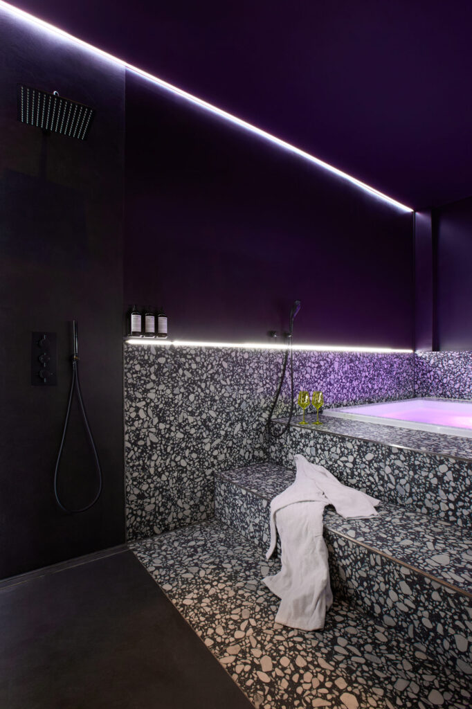 A wide angle view of the mini spa at The Circle Berlin, showcasing the heated steps with two wine glasses, a draped luxury bathrobe, atmospheric purple lighting, a rainfall shower, and organic L:A Bruket bodycare products.