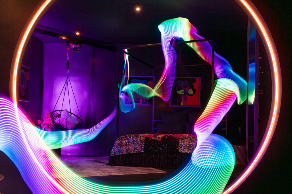 A view from the kitchen and lounge area looking into the bedroom suite at The Circle Berlin private spa, showcasing the custom-made kingsize 4-poster bed with a Tata Christiane sequined bedspread under colorful, whimsical lighting effects by Electric Skin.