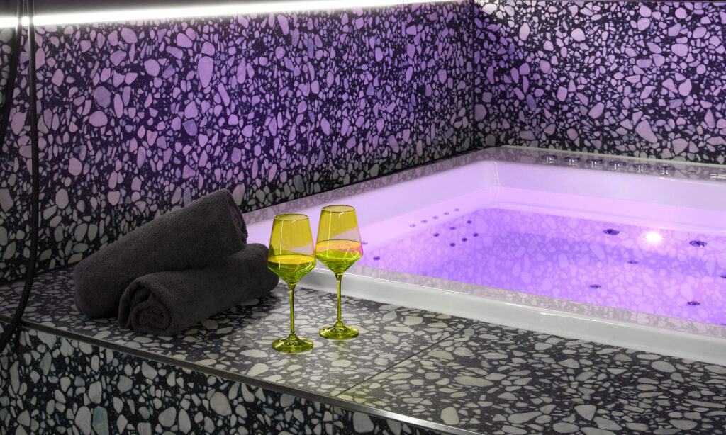 Two yellow wine glasses resting on heated steps leading to an 1.80x1.80 cm whirlpool jacuzzi with moody purple lighting, a womanizer showerhead, folded towels, and premium terrazzo tiles at The Circle Berlin private spa.