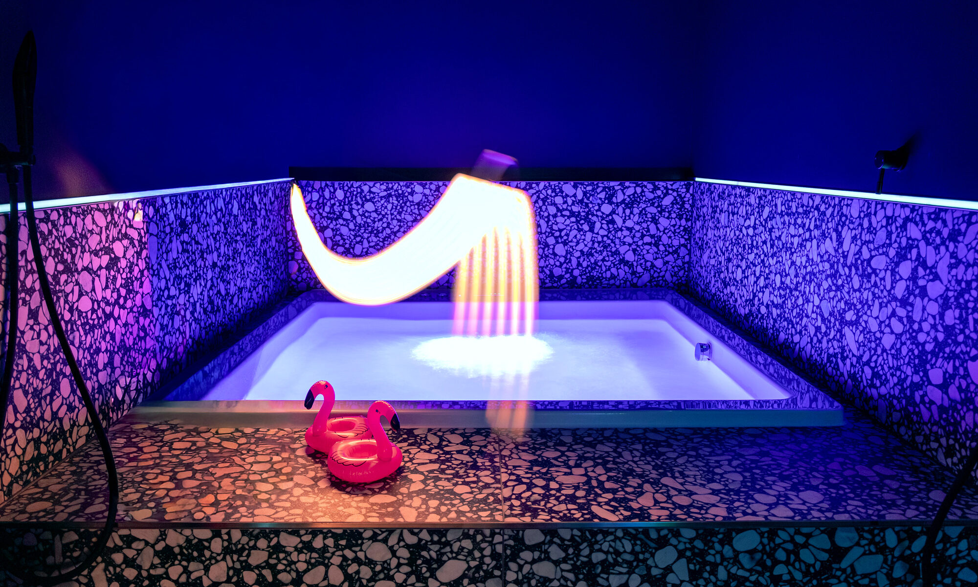 A view into the full jacuzzi at The Circle Berlin private spa, featuring two pink plastic flamingos on the heated top step under a colorful, whimsical lighting effect by Electric Skin.